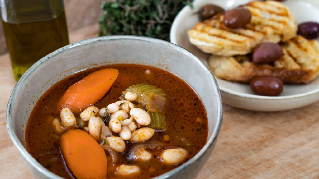 eine Schale mit griechischer Bohnensuppe, Brot und Oliven im Hintergrund