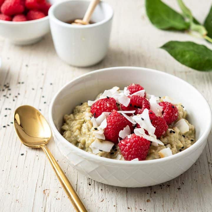 Image of white bowl with matcha oatmeal, topped with raspberries, on a white wood background, with leaves to the side