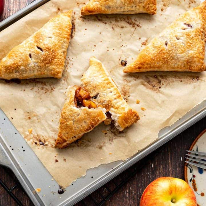 Four apple turnovers on a paper-lined baking sheet with apples