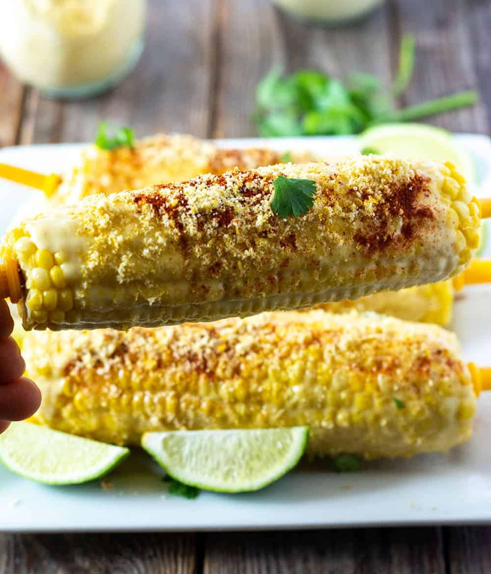 Mexican corn on a white platter, slathered with vegan mayo, vegan parmesan, cayenne pepper and lime juice