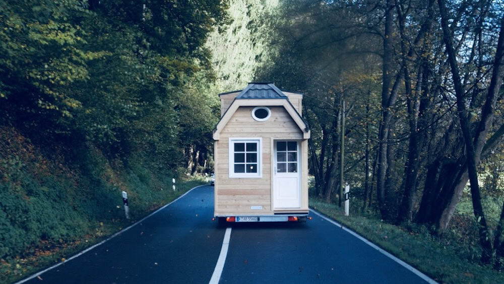 Bild zeigt den Transport eines Hauses auf Rädern. Zu sehen ist eine Straße und ein Wald. Das Haus auf Rädern wird auf einem Trailer von einem PKW gezogen