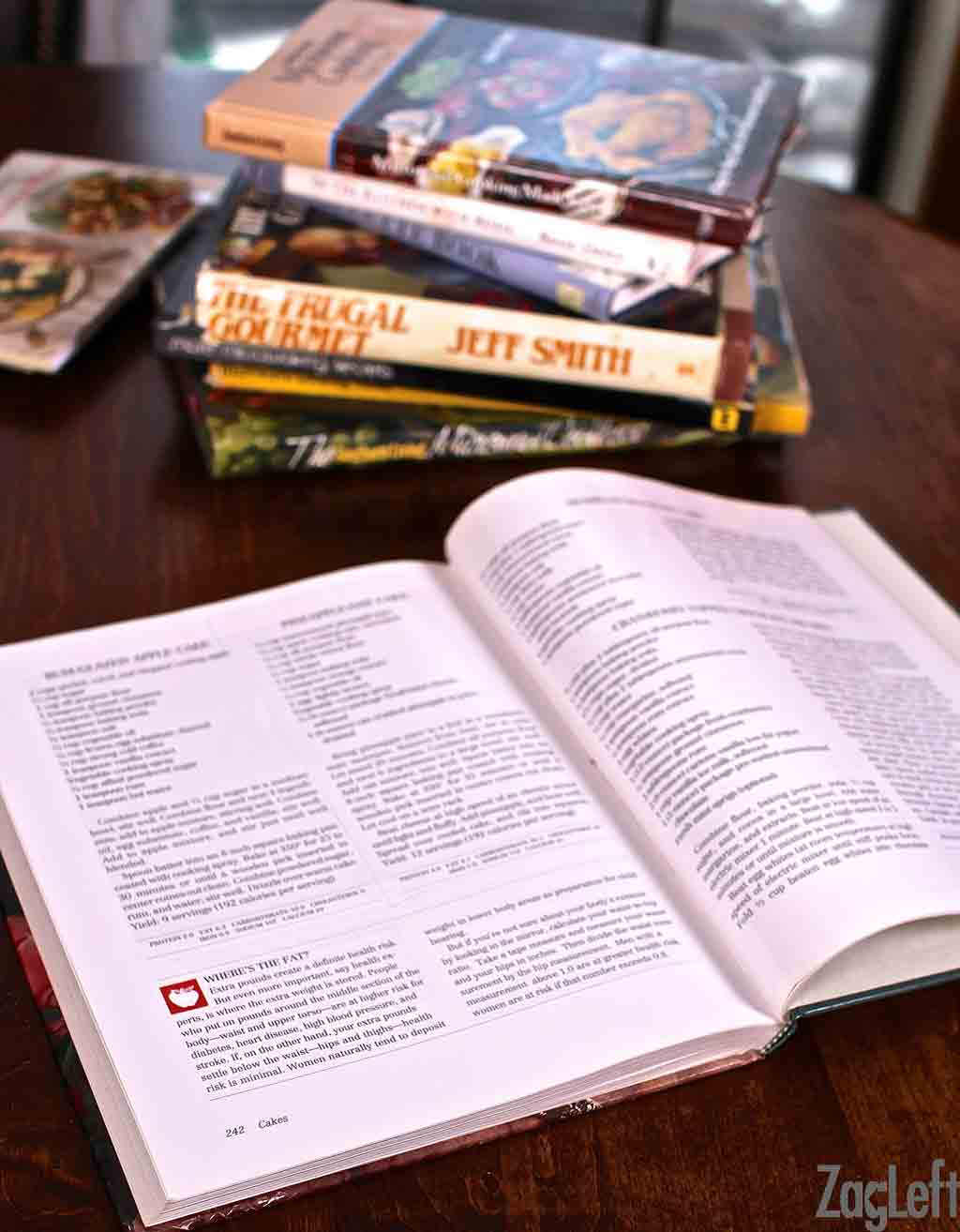 An open cookbook next to a stack of vintage cookbooks
