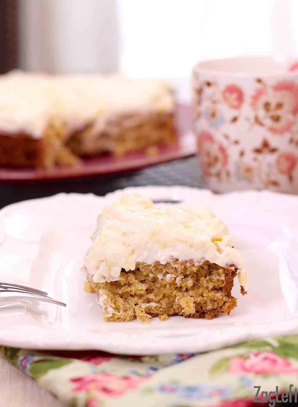 Closeup of a slice of pineapple oat cake with cream cheese frosting on a plate with a floral napkin and a coffee mug in the background