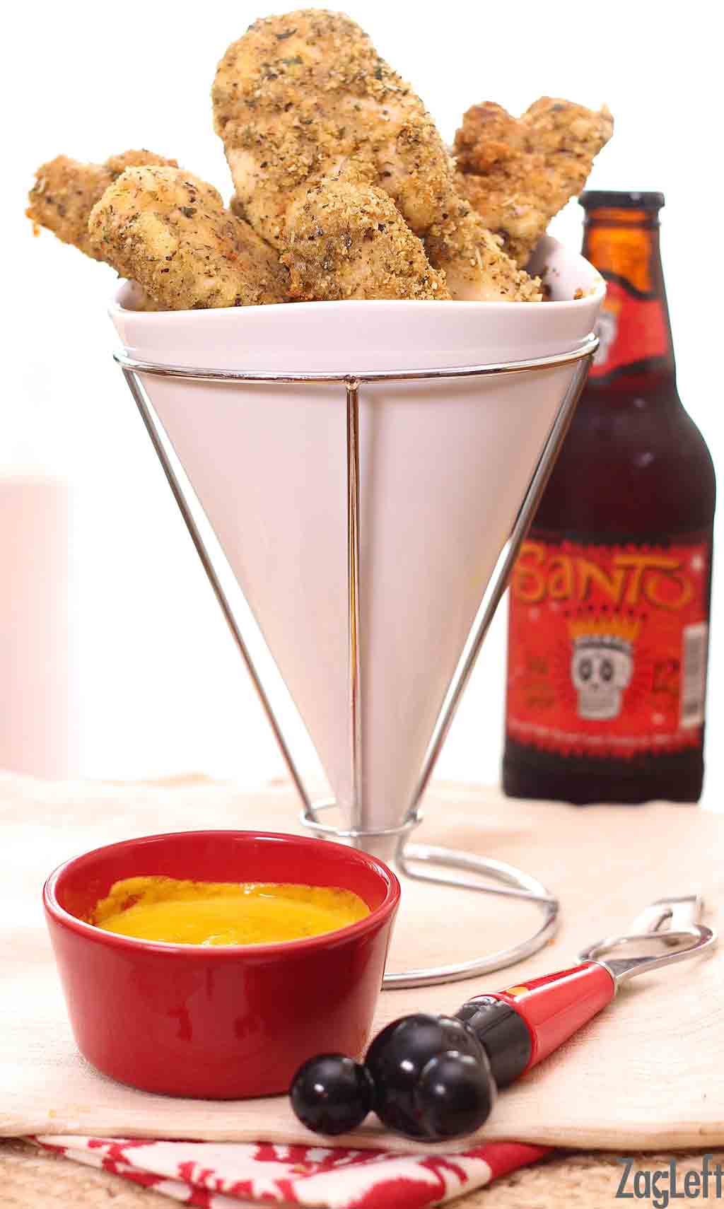 A small batch of chicken tenders baked in an oven arranged in a tall dish next to a bottle of beer and small bowl of honey mustard