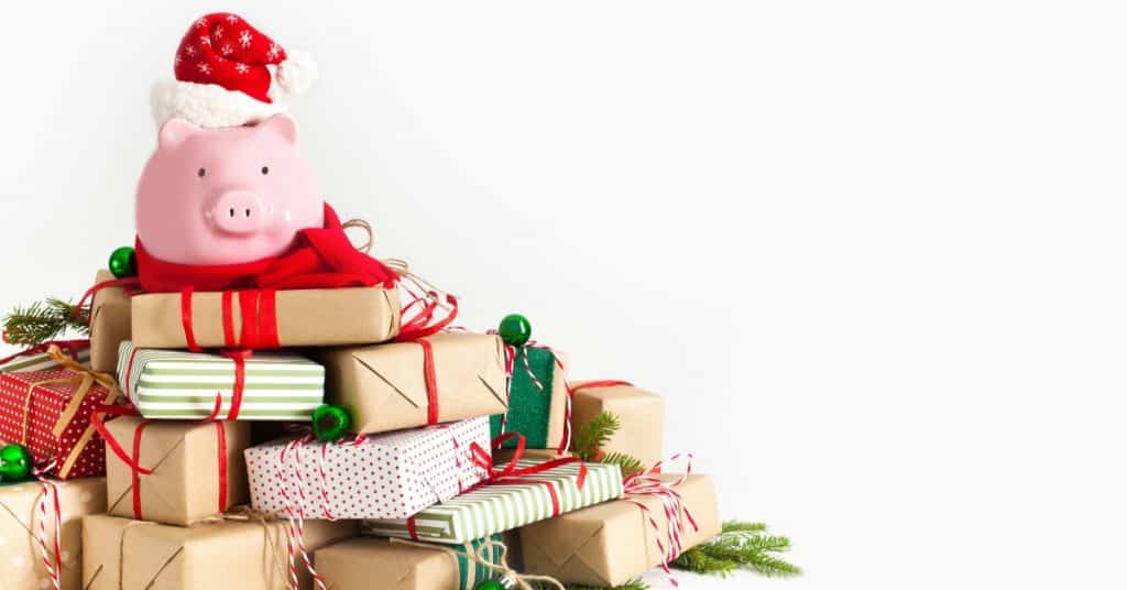 Piggy bank with Santa hat sitting on top of a large pile of Christmas presents and text overlay "How to stay on budget this holiday season"