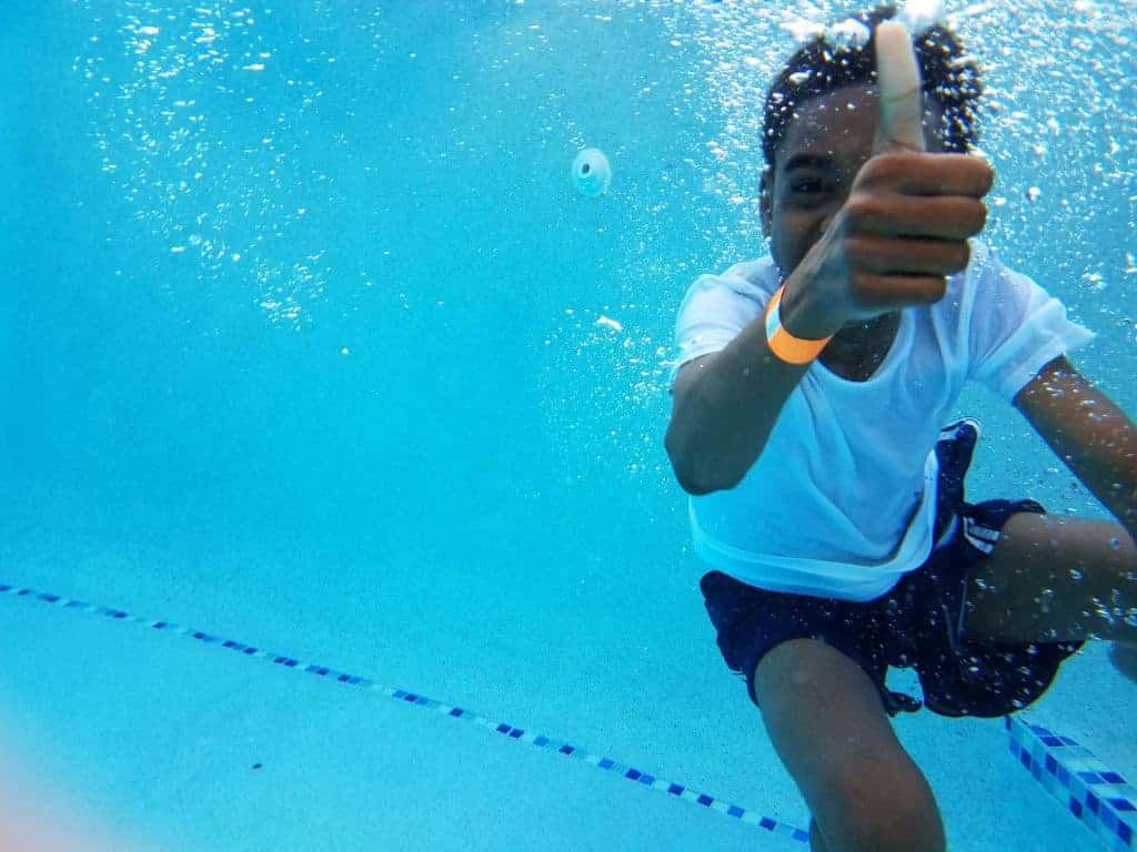 kid with white shirt in swimming pool under water doing a thumbs up