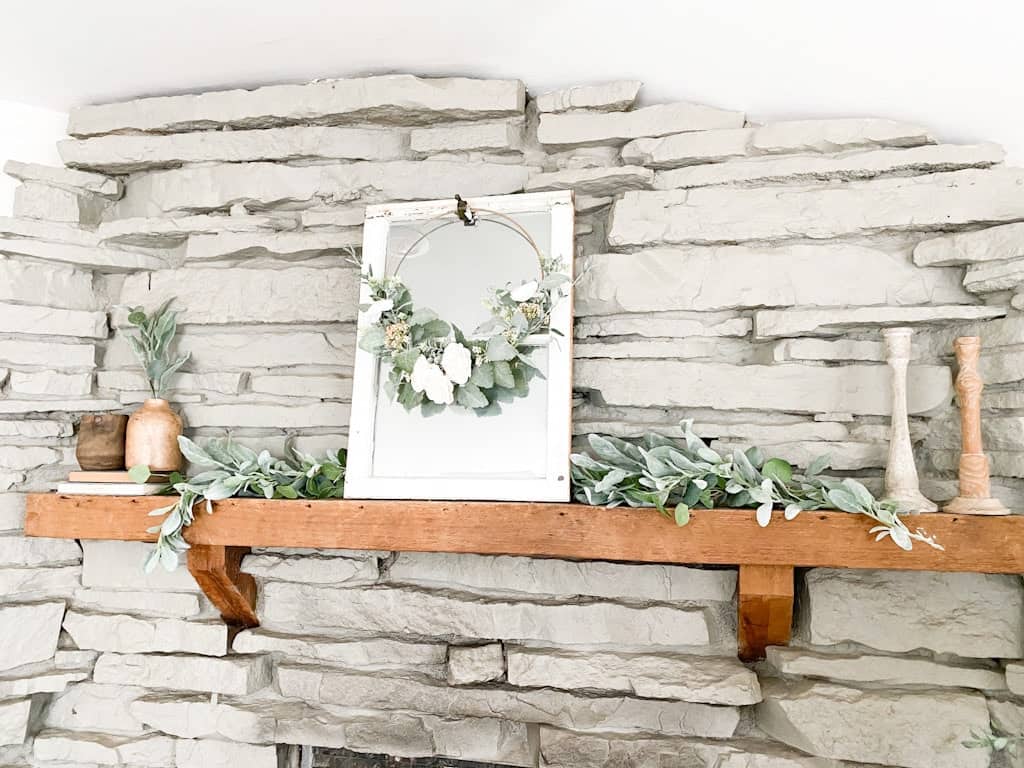 This image shows a gray stone fireplace with a wood mantel decorated with a garland, a window mirror, a spring hoop wreath, some wooden candle holders, and some wooden vases on top of old books.