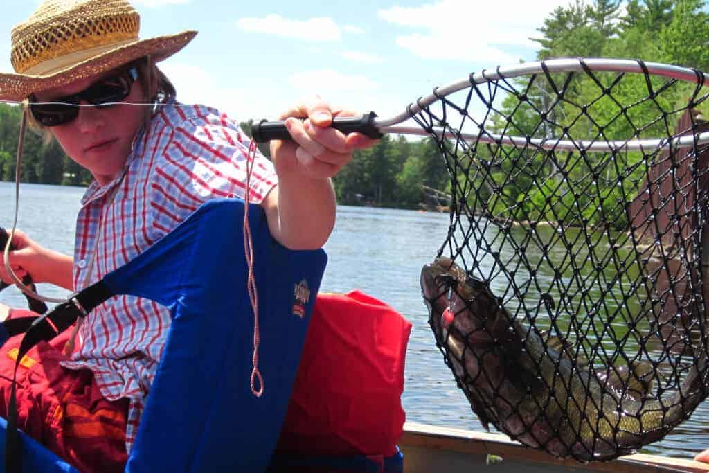 A boy sits in a canoe with a netted fish. He is wearing sunglasses and a straw hat.