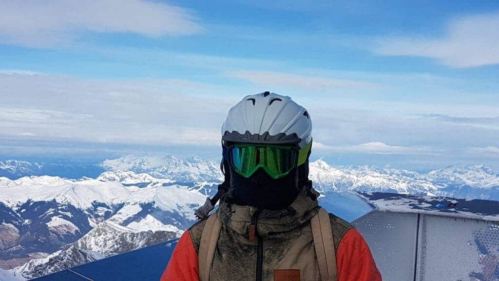 family ski vacations A person all decked out in ski gear stands at the top of a mountain with snow-capped peaks in the background.