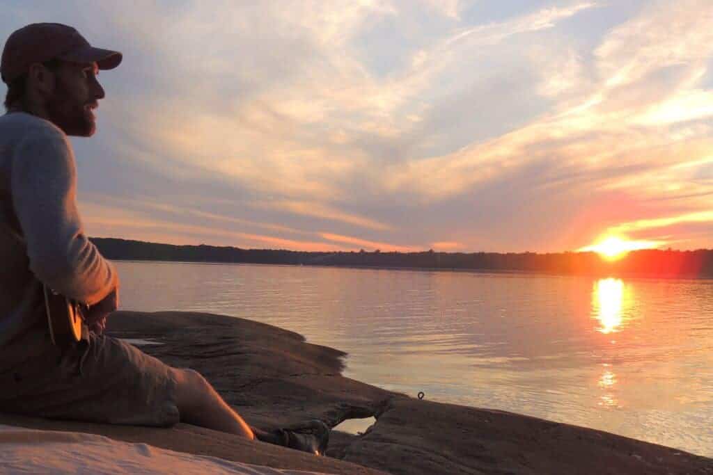 Why you should road trip as a couple A man sits facing the setting sun, playing the guitar on a couples road trip.