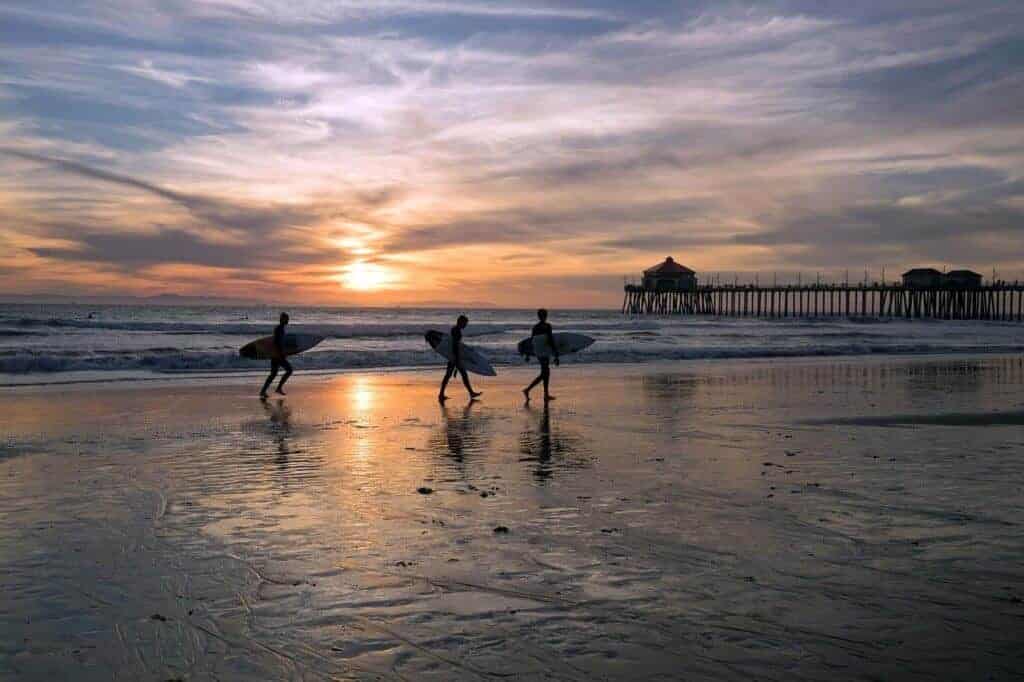 A group of three surfers walk along the shore during sunset at Huntington Beach, CA.