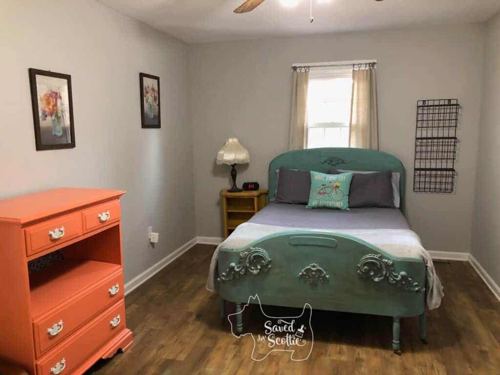 The Goat Loft bedroom with grey walls, ornate wooden bed painted teal, coral dresser, mustard yellow nightstand with lamp and clock on it. Tiered basket rack next to bed on right, window with curtains behind bed and two floral prints on the left wall that tie all the colors together.