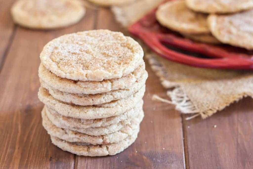 stack of gluten free snickerdoodles with cinnamon sugar on a wooden table with red tray of cookies in the bacground