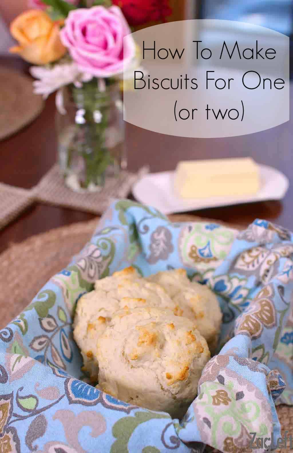 Three biscuits in a small basket lined with a blue cloth napkin next to a stick of butter on a plate and a small vase of flowers