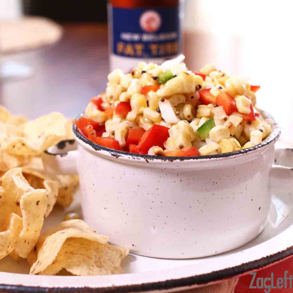 A bowl of Corn Salsa plated with tortilla chips next to a bottle of beer