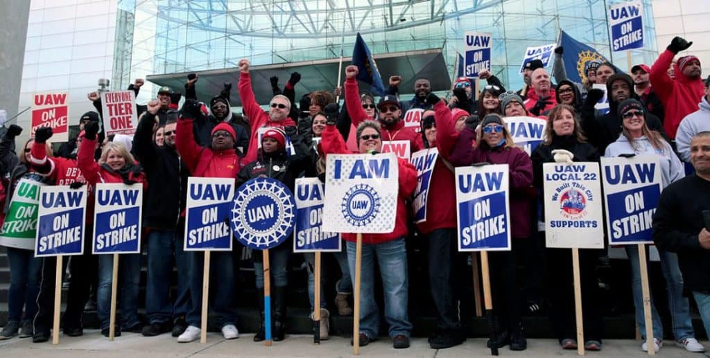 A large multiracial group of men and women with UAW picket signs.