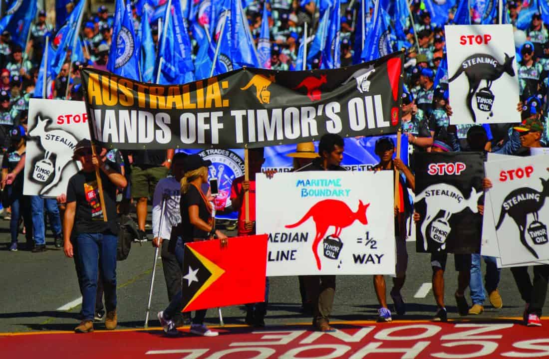 People marching with a large banner that reads AUSTRALIA: HANDS OFF TIMOR OIL. Another sign has a kangaroo graphic and the text MEDIAN LINE = 1/2 WAY. referencing the median line.