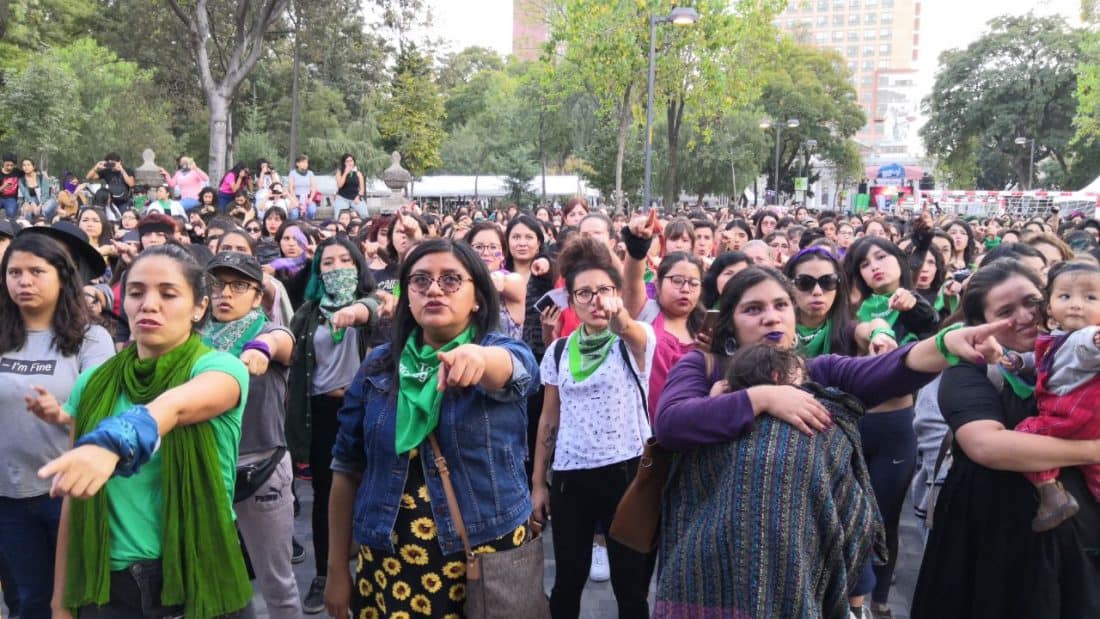 A crowd of women, some carrying babies, in a park gesture toward the camera.