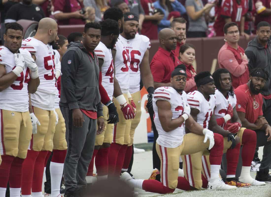 Football players on the sidelines, some standing and some kneeling. They are giving each other questioning looks.