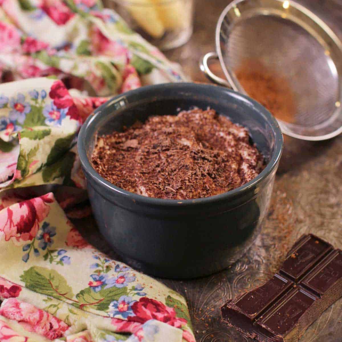 A mini tiramisu topped with chocolate shavings in a circular dish on a metal tray with a floral cloth napkin, a piece of a chocolate bar, and cocoa powder in a sieve
