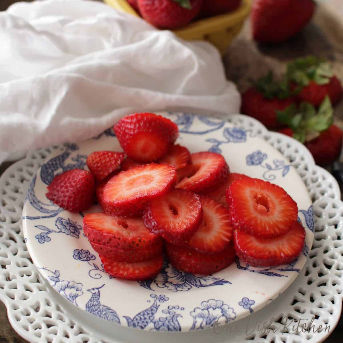 A plate of fresh sliced strawberries next to a white napkin and a yellow bowl of whole strawberries.