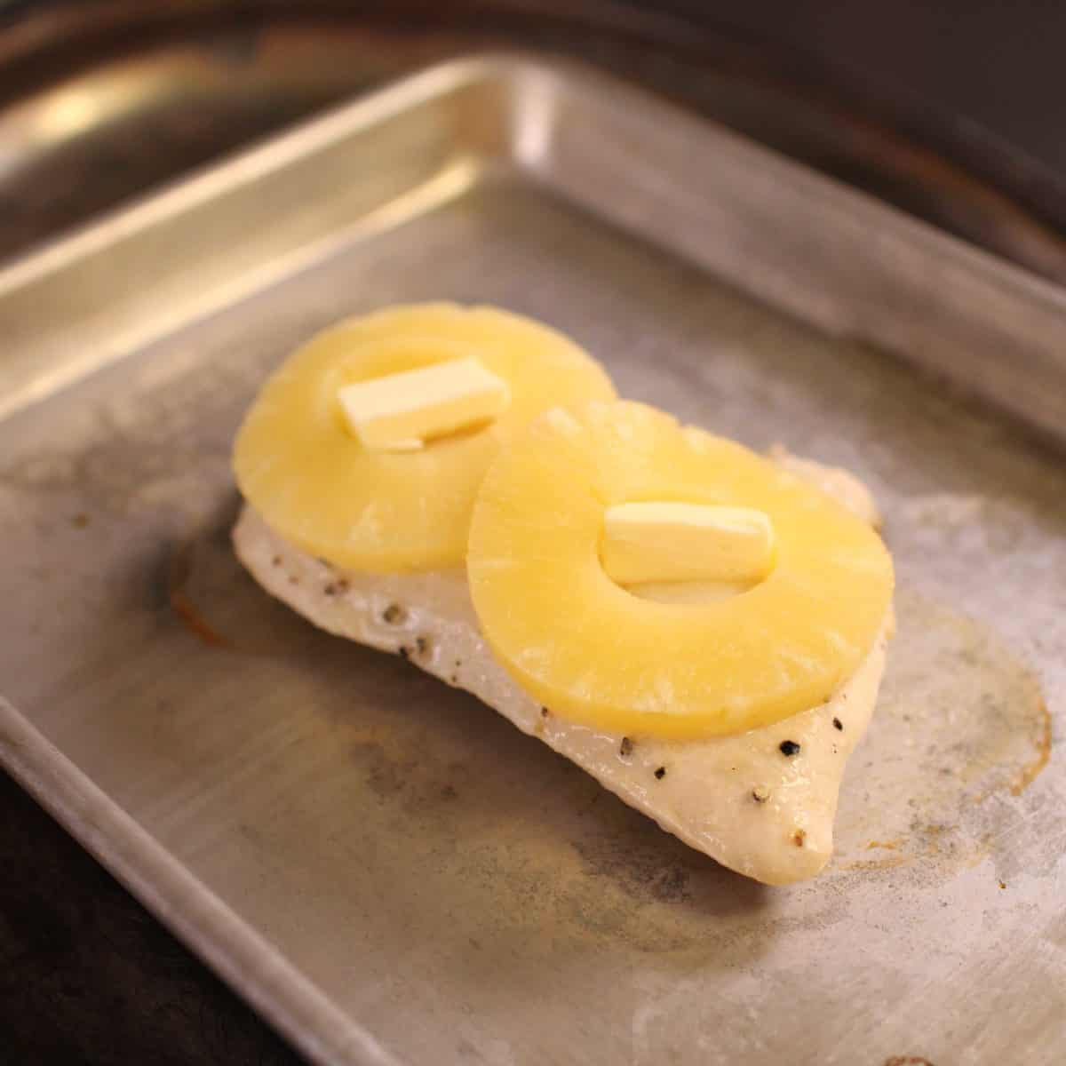 a piece of chicken on a baking sheet topped with butter and two pineapple slices.