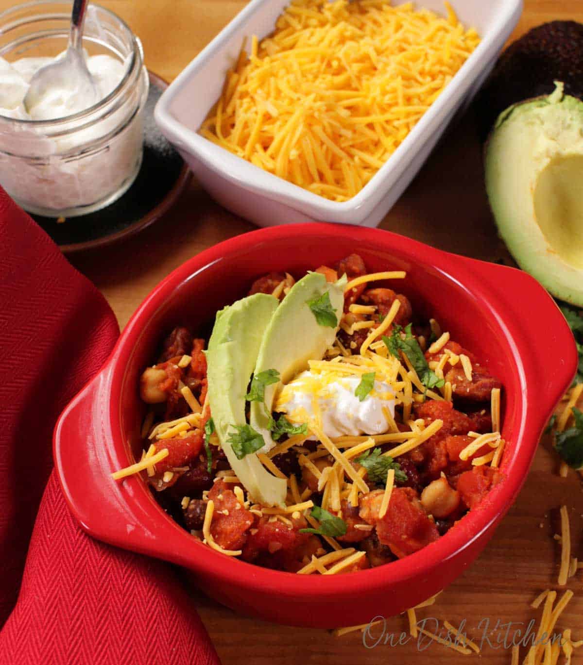 An overhead view of a circular baking dish of vegetarian chili topped with two slices of avocados, shredded cheese, and a dollop of sour cream next to a small jar of sour cream, shredded cheese, and half of an avocado
