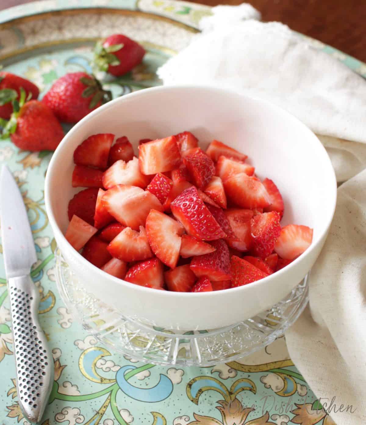 A bowl of fresh sliced strawberries plated next to a knife and scattered strawberries