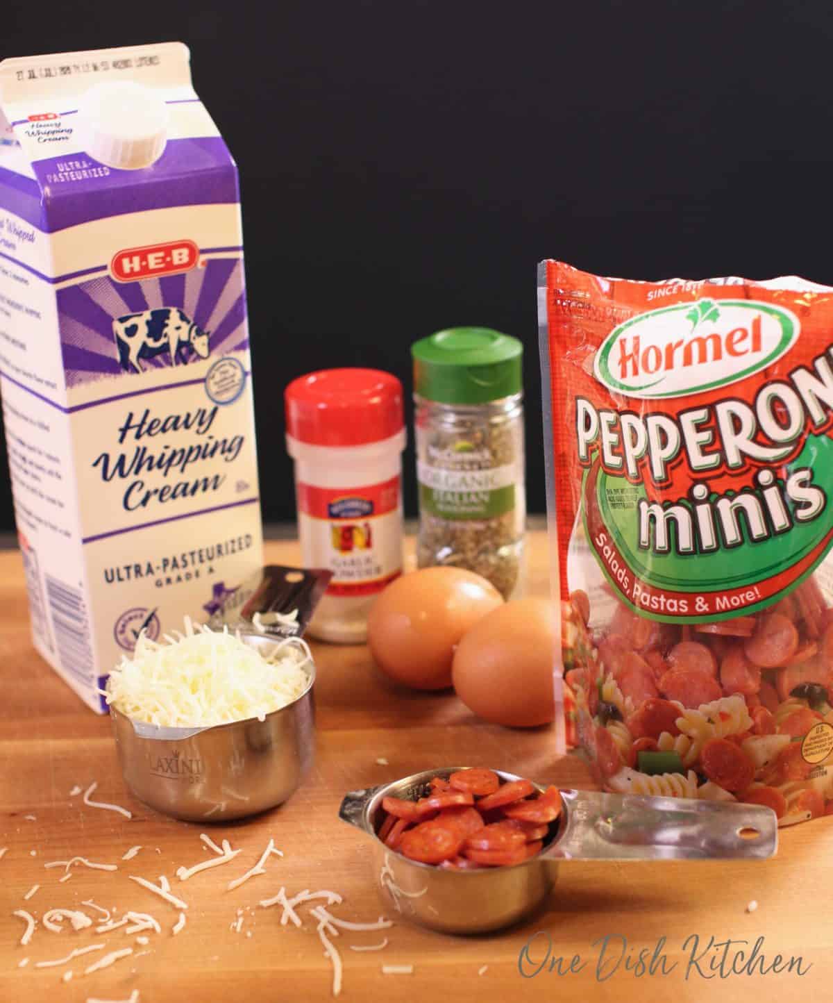 a bag of pepperoni slices, a measuring cup filled with shredded mozzarella cheese, a carton of heavy cream and spices on a brown table.