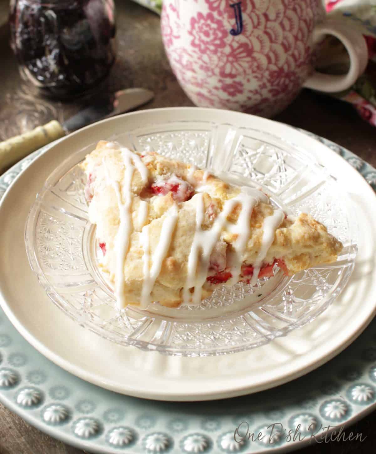 A scone on a plate with drizzled white chocolate next to a cup of coffee and a small jar of jam