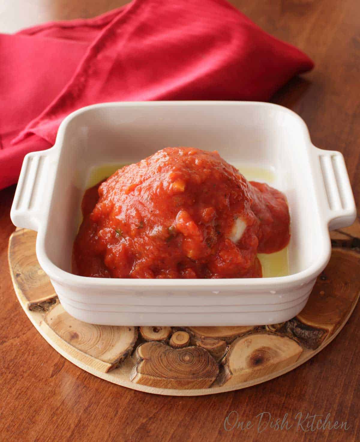 A meatball topped with tomato sauce in a small baking dish on a wooden trivet