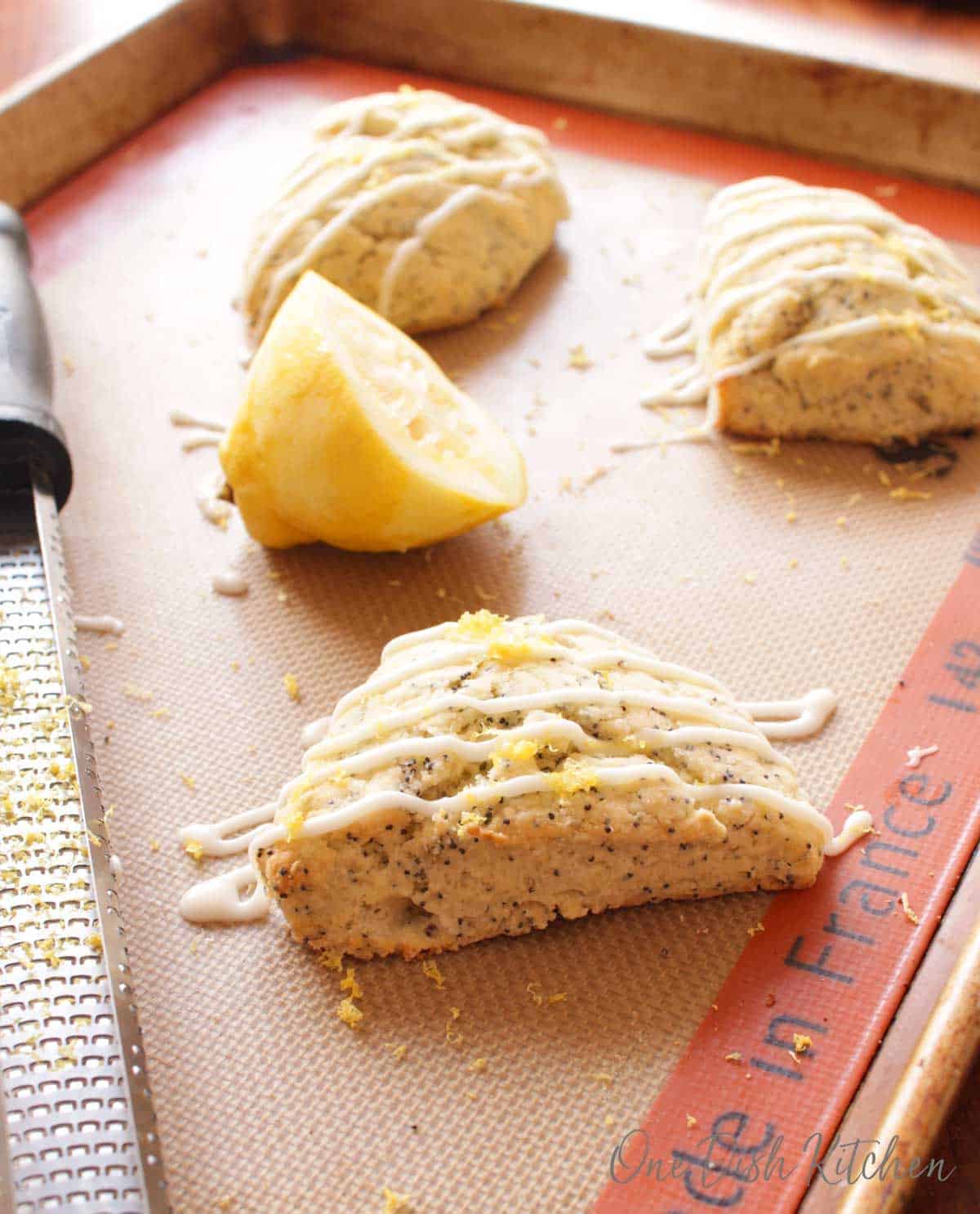 Three scones topped with a white glaze and lemon zest on a baking sheet next to a half of a lemon.