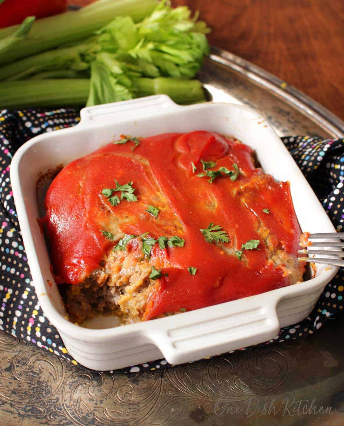A forkful of bacon cheeseburger meatloaf missing from a small baking dish on a metal tray next to a bunch of celery