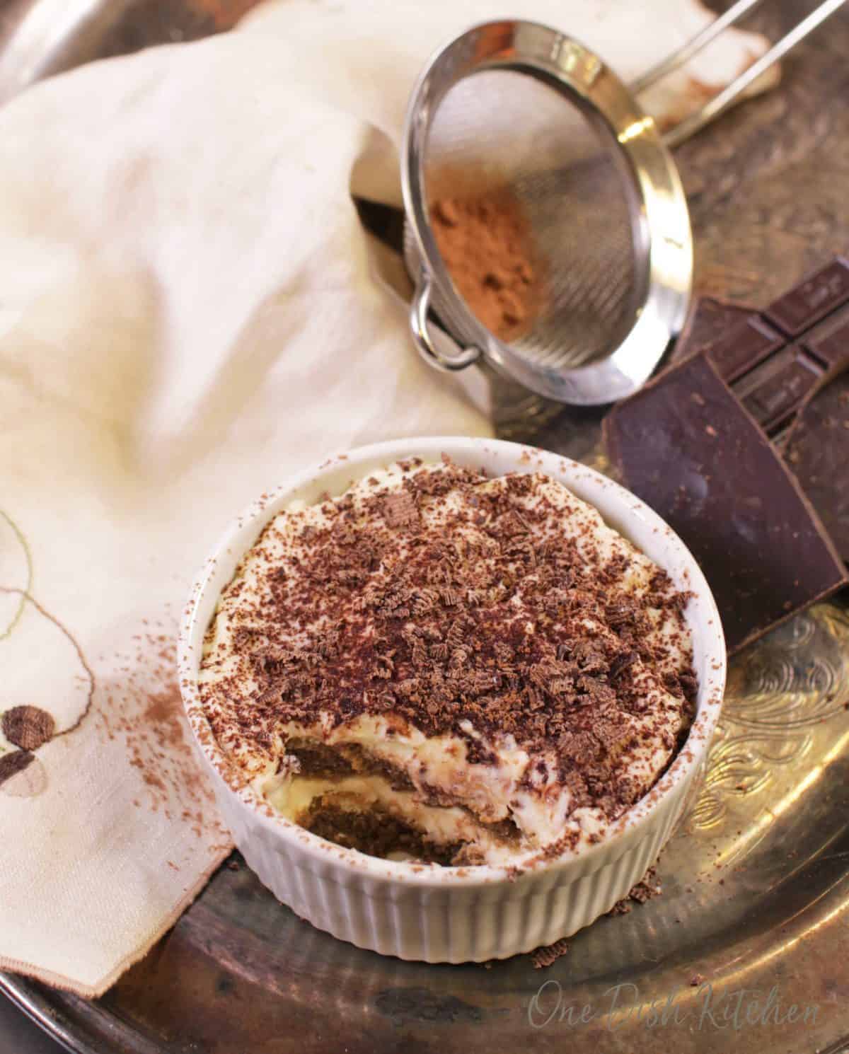 A mini tiramisu topped with chocolate shavings in a ramekin next to a piece of a chocolate bar, a cream-colored cloth napkin, and cocoa powder in a sieve all on a metal tray