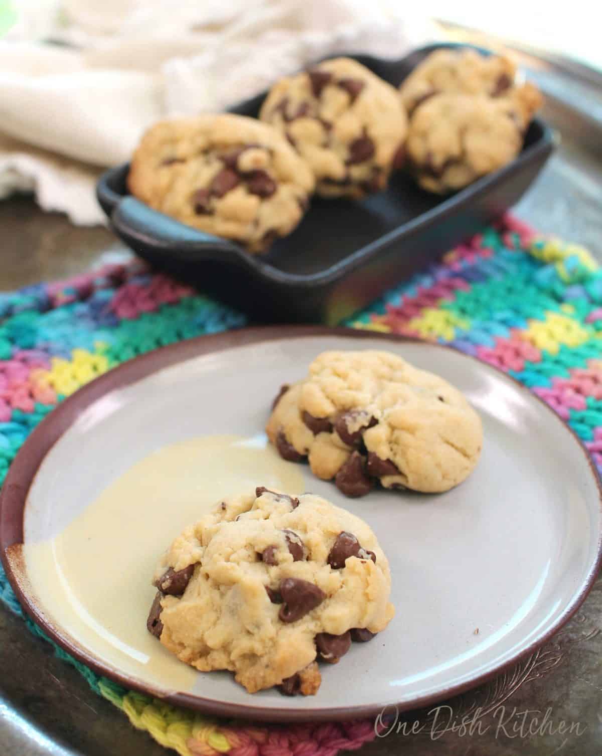 two chocolate chip cookies on a blue plate with a bowl of cookies in the background.