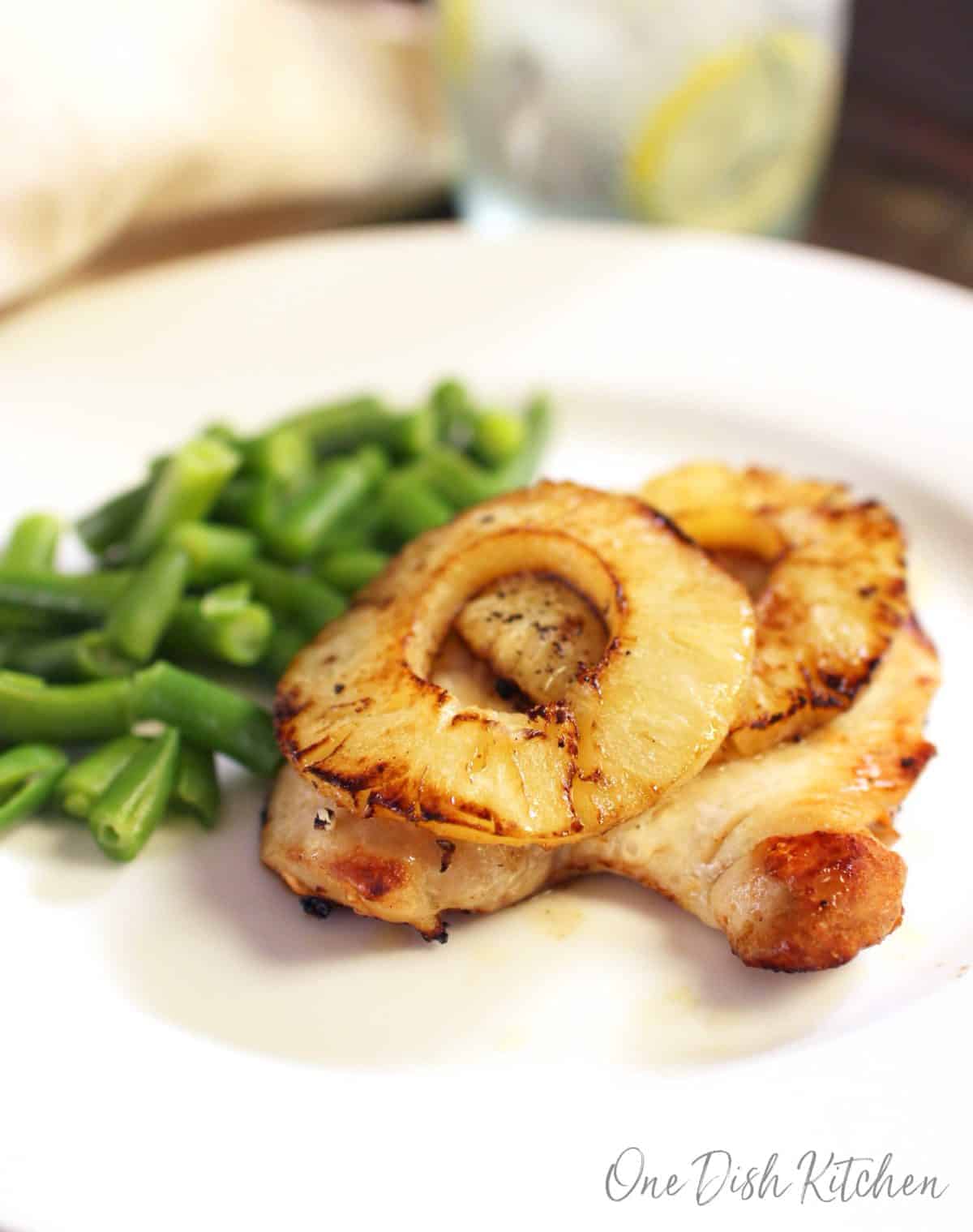 one chicken breast on a white plate with slices of pineapples on top next to cooked green beans.