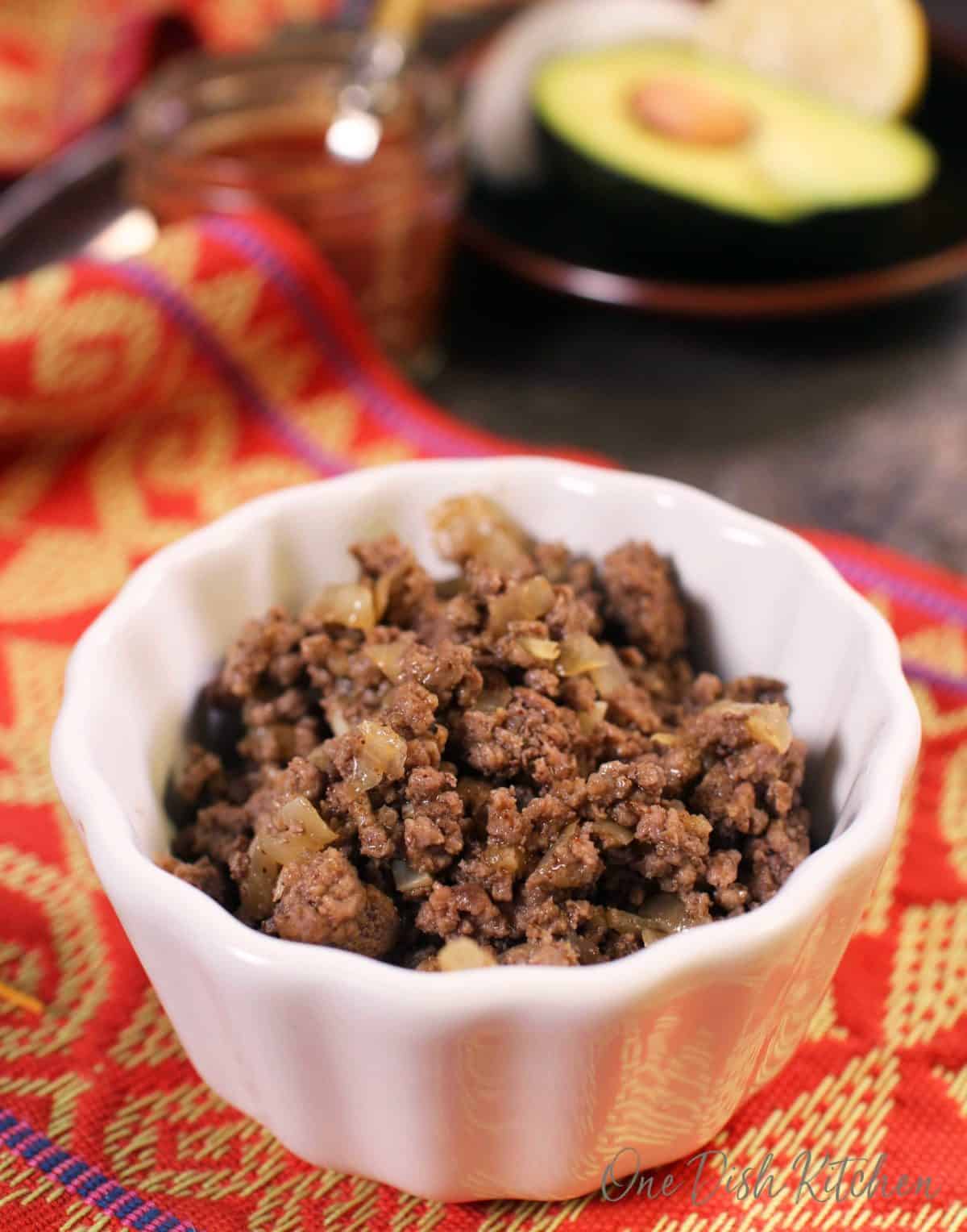 A closeup of a small bowl of ground beef and cooked onions with half of an avocado in the background