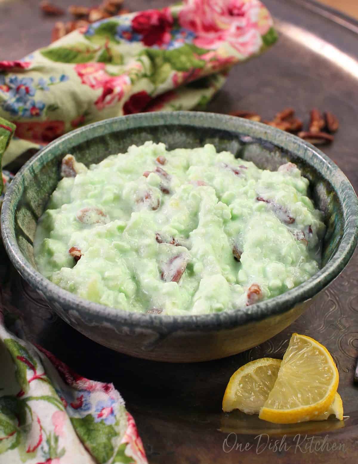 a blue bowl filled with green jello fluff topped with nuts on a silver tray next to two lemon slices.