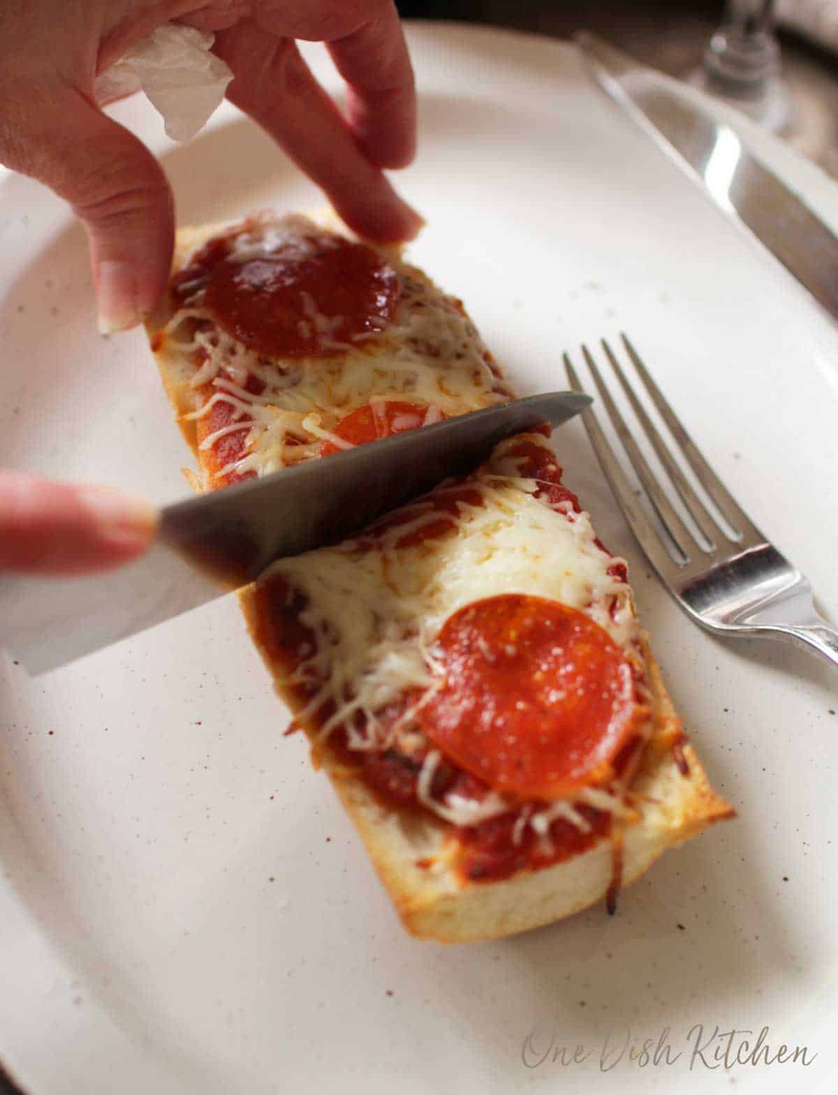 A knife cutting into french bread pizza that is topped with mozzarella cheese and pepperoni slices