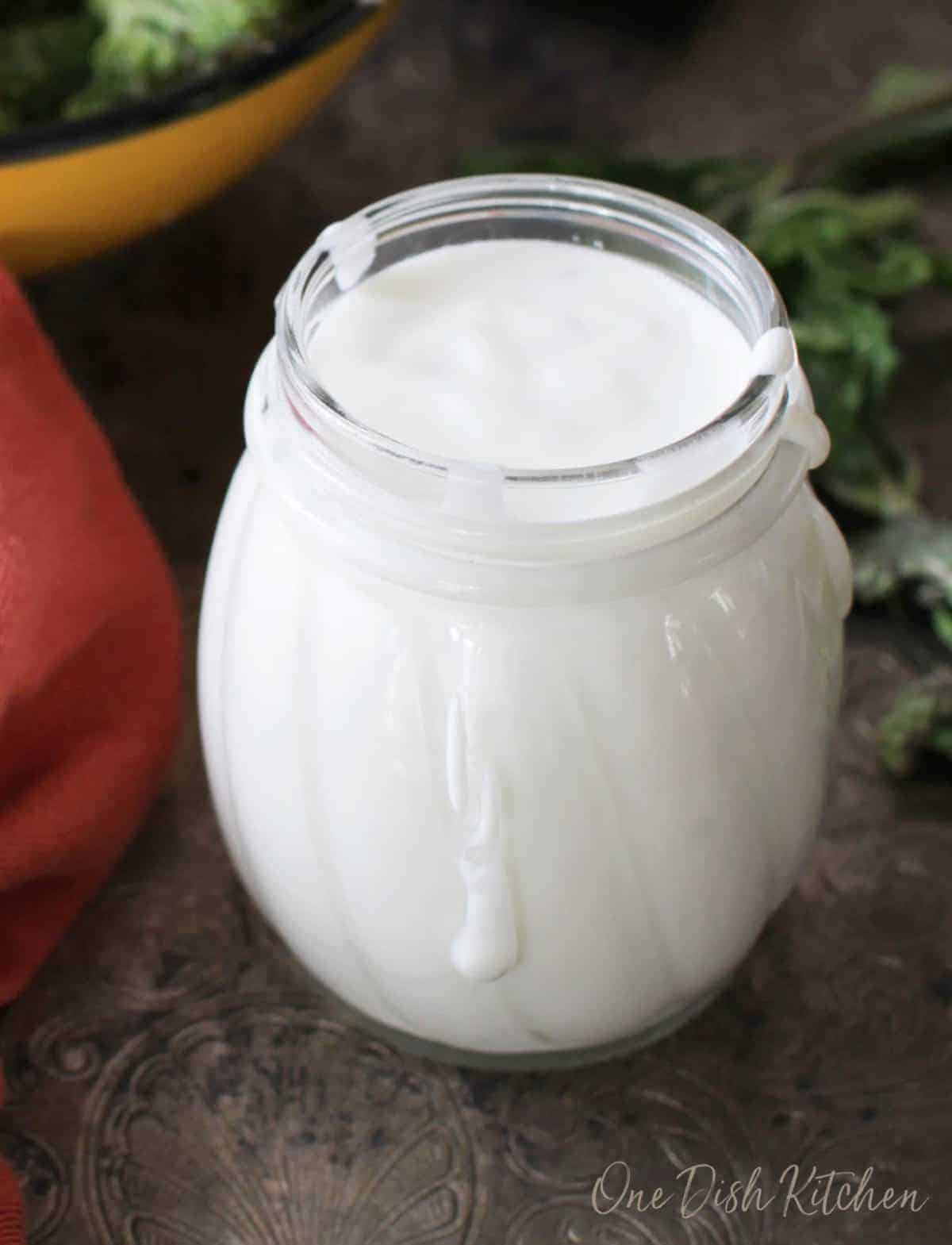 A closeup of a small jar of goat cheese salad dressing on a metal tray with drips of dressing around the sides.