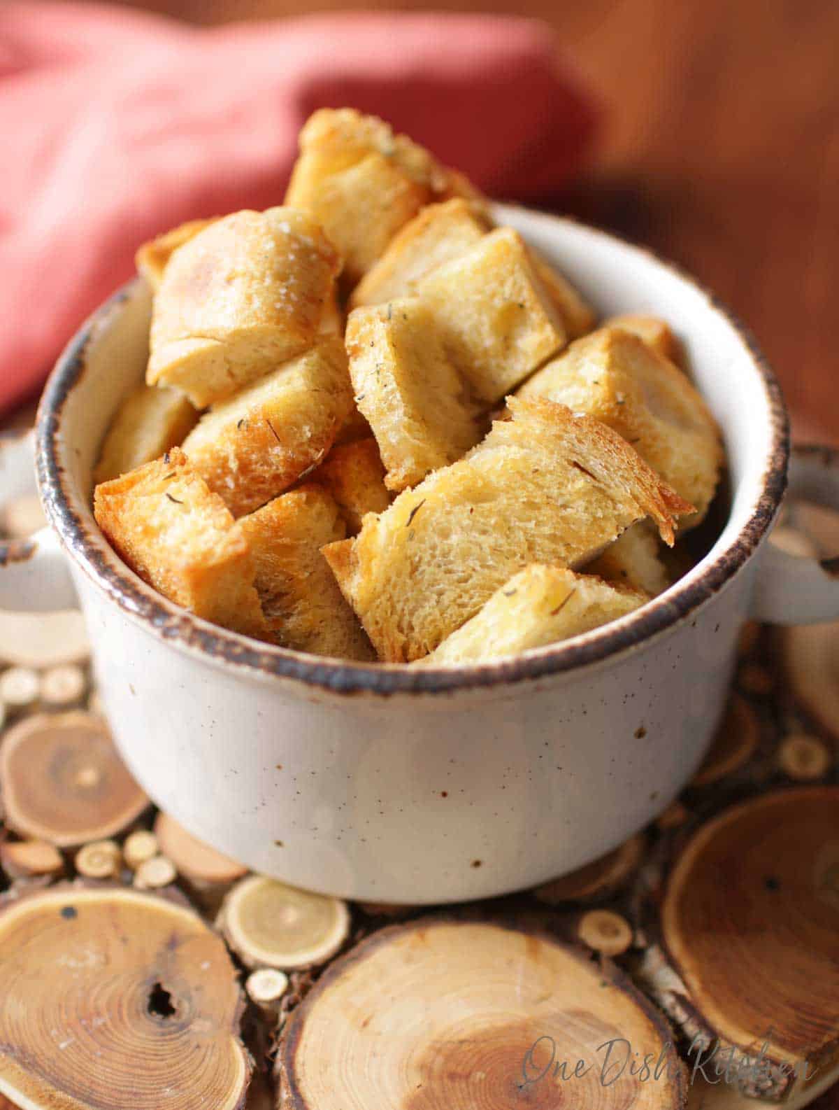 A closeup of a bowl of baked croutons on a wooden trivet