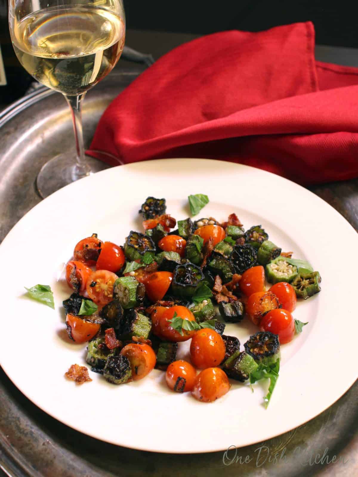 Fried okra, bacon, and tomatoes plated on a metal tray with a glass of white wine and a red cloth napkin