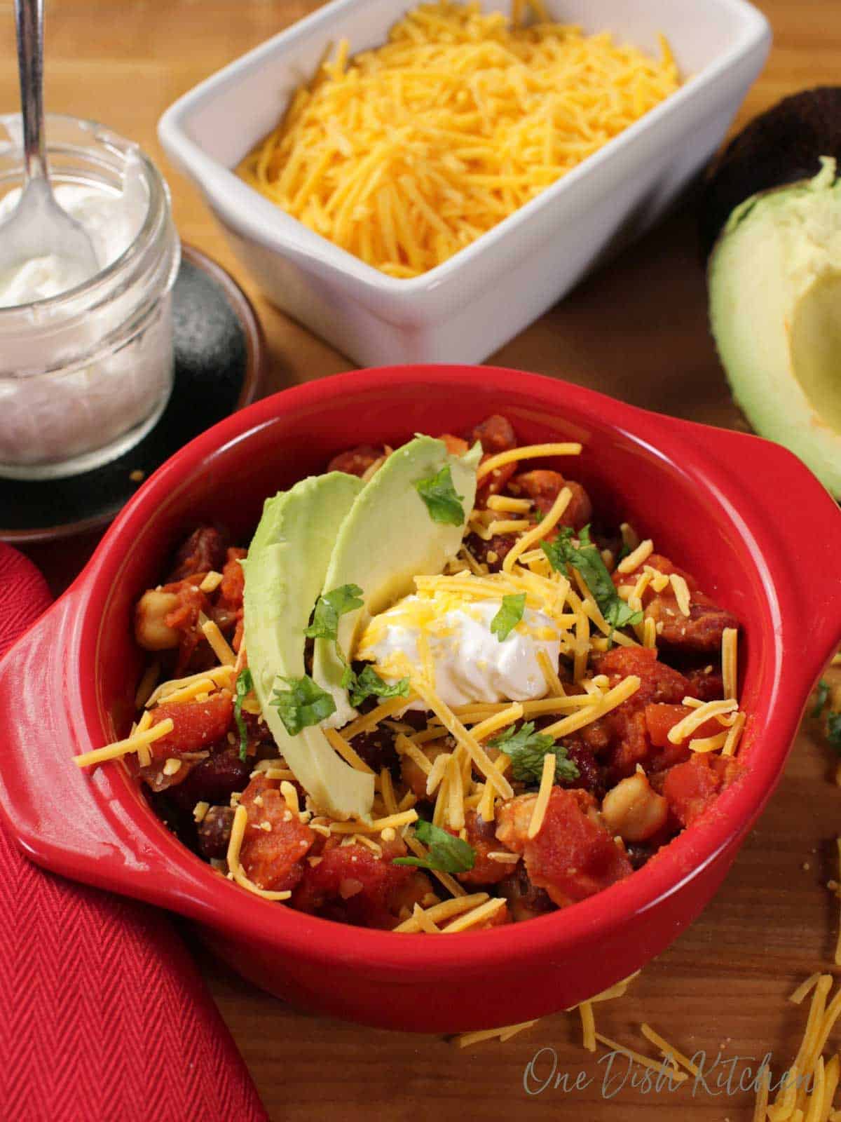 A red circular baking dish full of chili with beans, onions, and tomatoes, topped with two avocado slices, shredded cheese, and a dollop of sour cream, next to a small jar of sour cream, a dish of shredded cheese, and half of an avocado