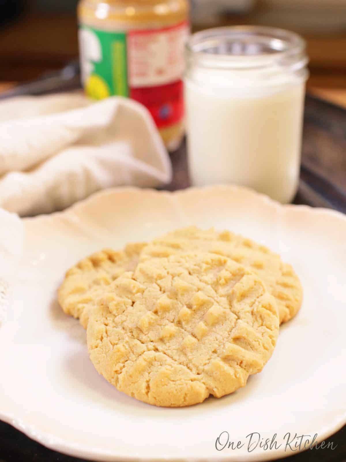 Three peanut butter cookies on a plate next to a jar of peanut butter and a glass of milk.