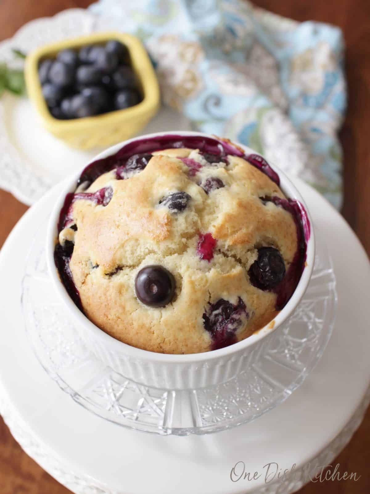 An overhead view of a blueberry muffin in a ramekin plated next to a small bowl of blueberries