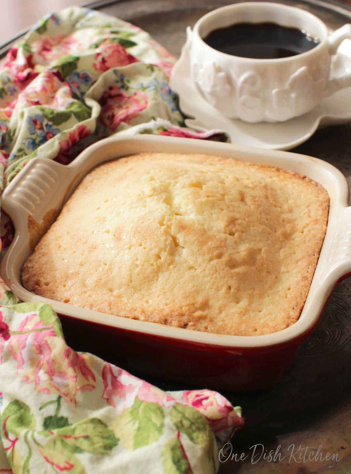 One square pound cake in a small baking dish next to a cup of coffee.