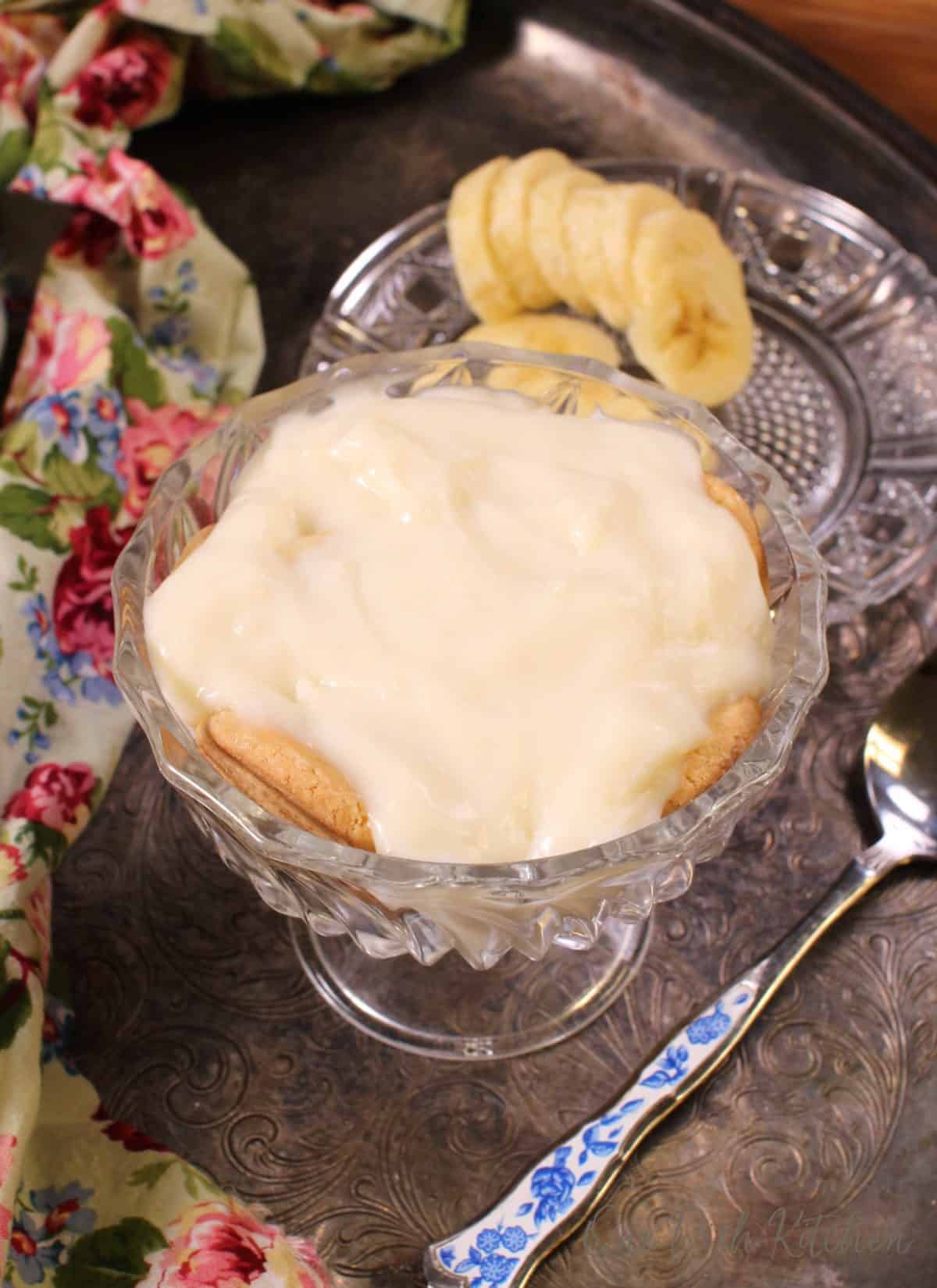 An overhead view of vanilla pudding on a bed of cookies and bananas in a dessert glass next to a floral cloth napkin, a small plate of sliced bananas, and a spoon