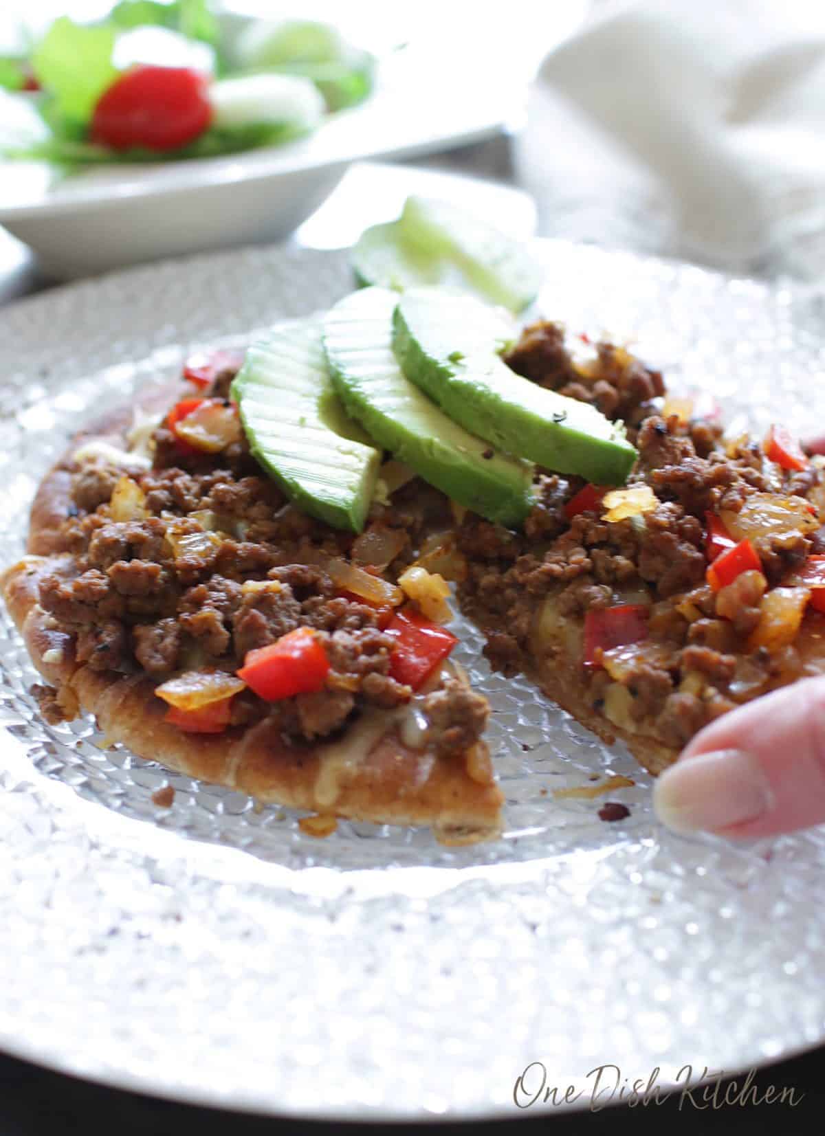 Hand grabbing a slice of pita pizza topped with ground beef, cheese, red peppers, onions, and three avocado slices next to a salad