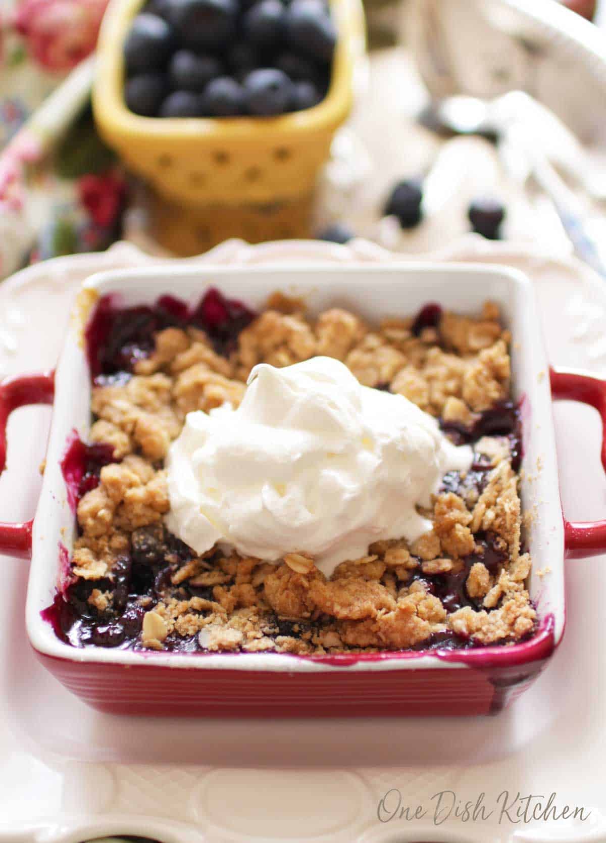Mini blueberry crumble in a small red baking dish topped with whipped cream next to a small bowl of blueberries all on a metal tray