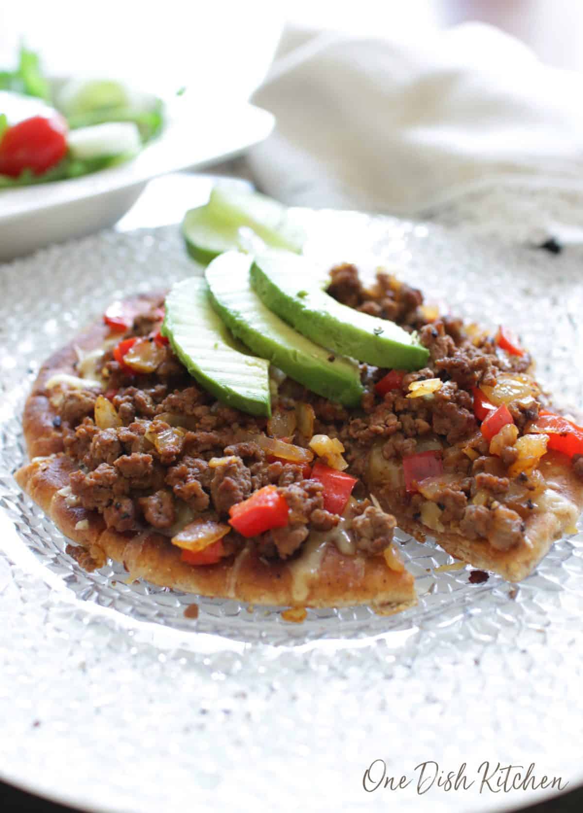 A pizza made with pita bread topped with ground beef, cheese, red peppers, onions, and three avocado slices
